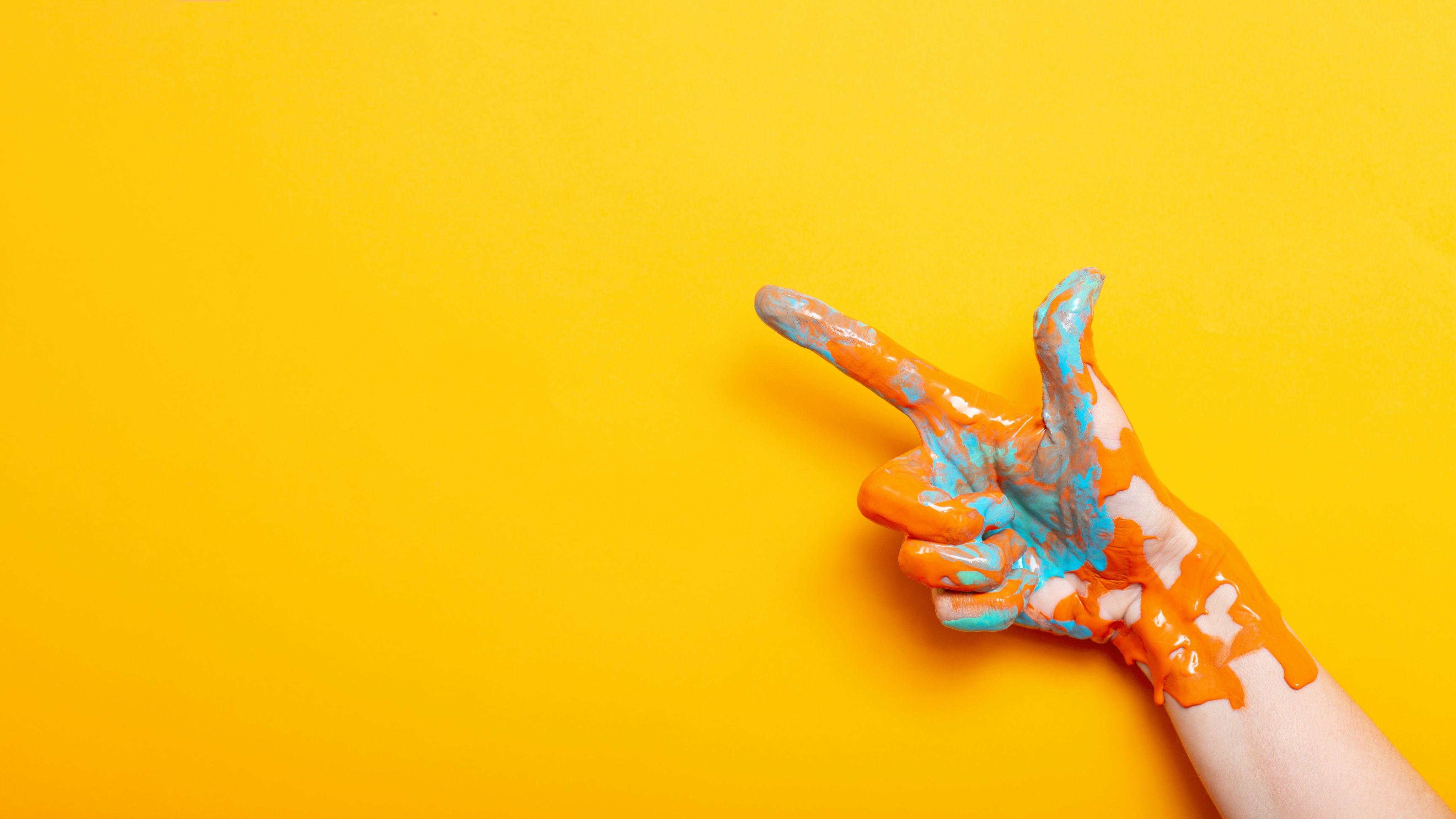 Hand covered in orange and teal paint against a yellow background pointing into the top left corner