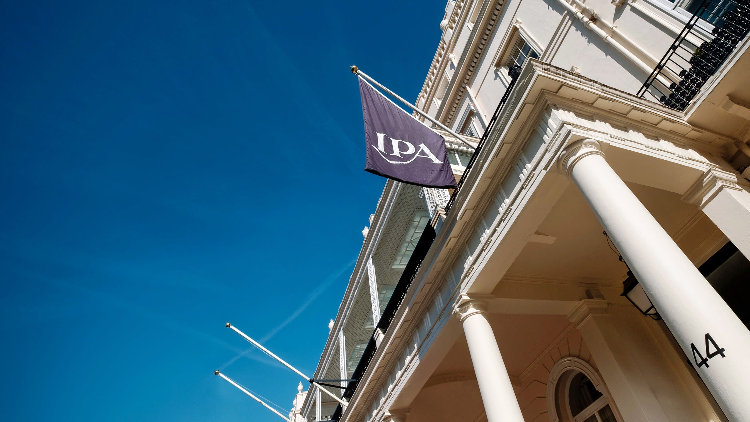 Low-angle view of a white neoclassical building with tall columns and a dark flag displaying the letters ‘IPA’ flying against a bright blue sky.