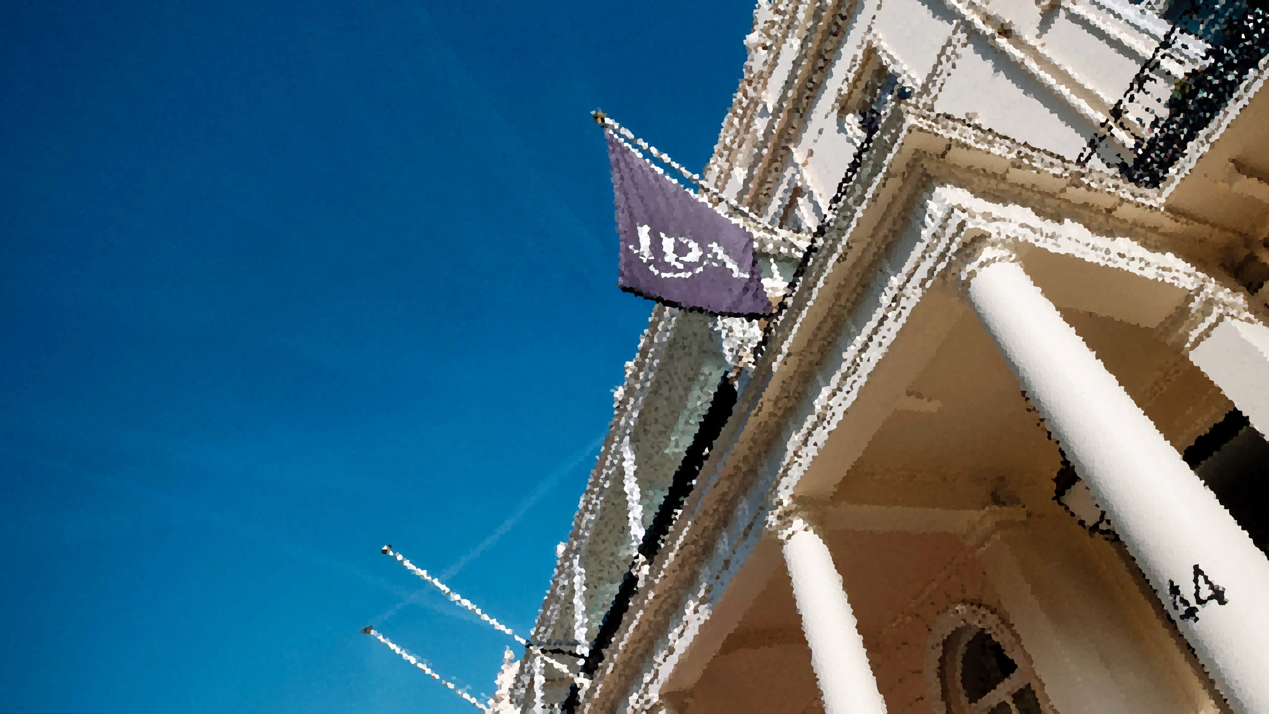 Low-angle view of a white neoclassical building with tall columns and a dark flag displaying the letters ‘IPA’ flying against a bright blue sky.